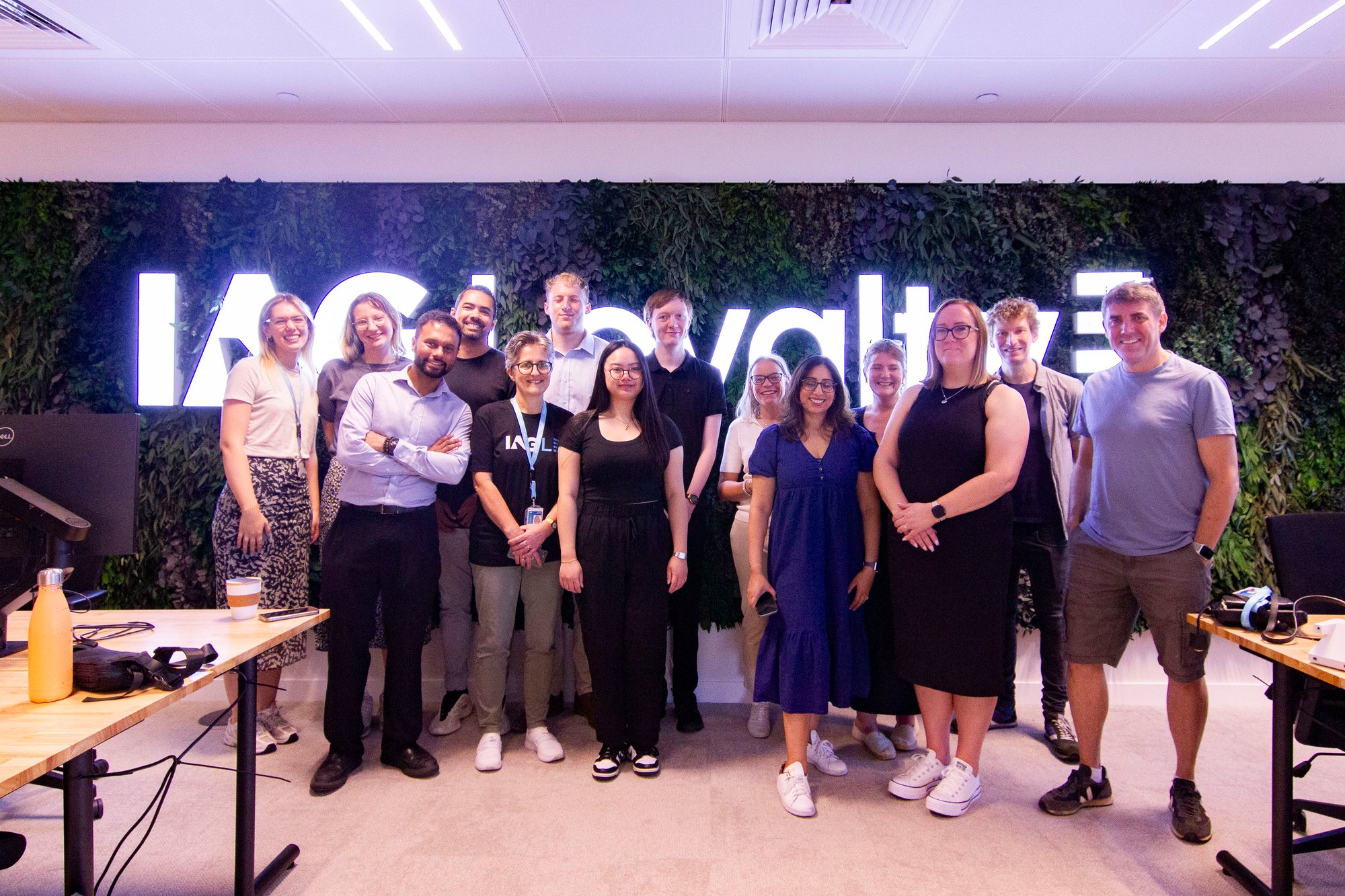 A group of colleagues smile in front of a neon plant wall that says IAG Loyalty A group of colleagues smile in front of a neon plant wall that says IAG Loyalty